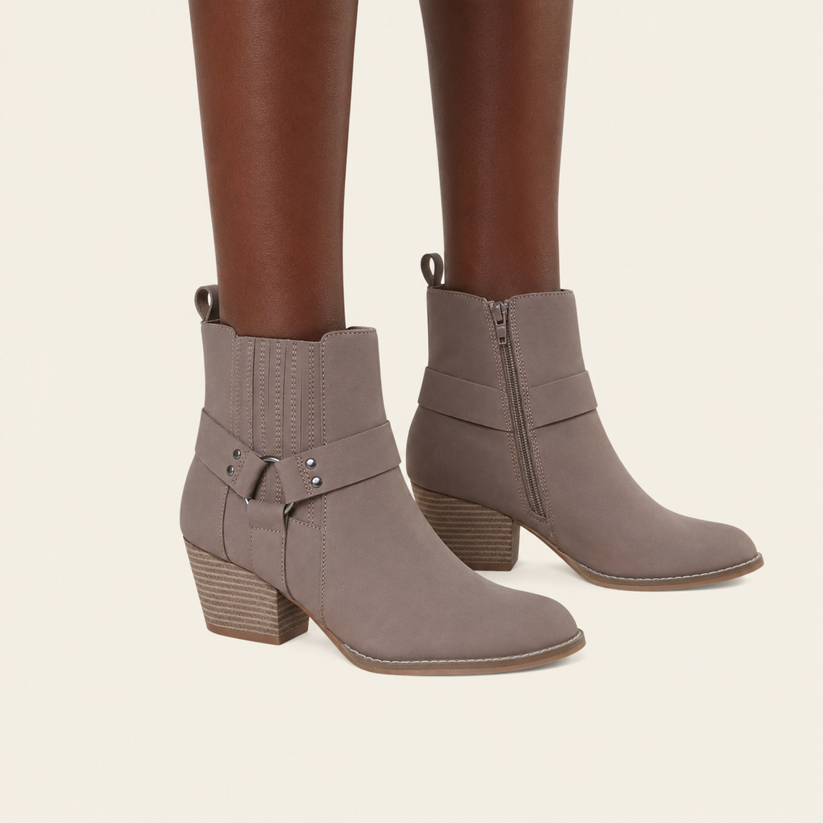 Taupe faux-suede ankle boots with side zippers and decorative buckle straps, shown on a model's feet against a light cream background.