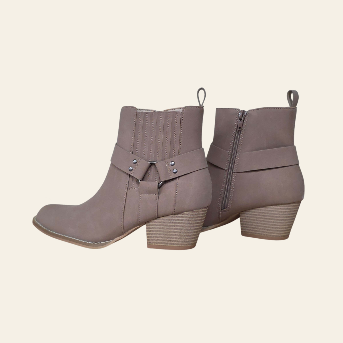 A pair of taupe faux suede ankle boots with a side zipper and a decorative strap with silver studs on the front, shown against a light cream background.
