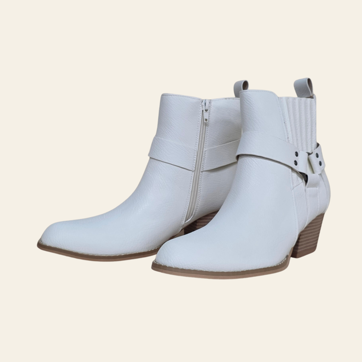 A pair of white faux-leather ankle boots with a side zipper, a decorative strap with a silver buckle on the outer ankle, and a stacked brown block heel, shown against a solid cream background.