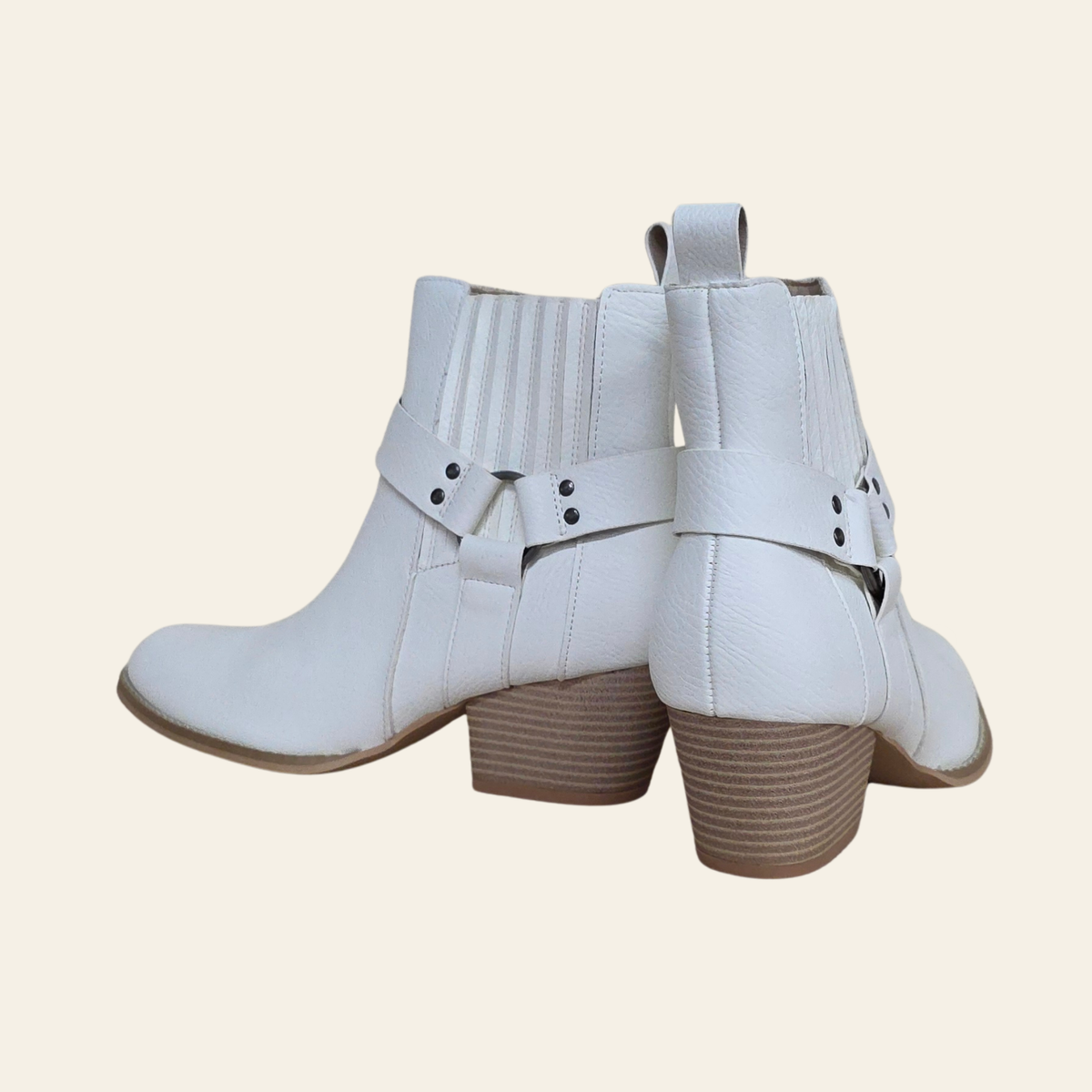 A pair of white faux-leather ankle boots with brown block heels, featuring decorative straps with silver-tone rivets and a pull tab, shown against a solid light beige background.