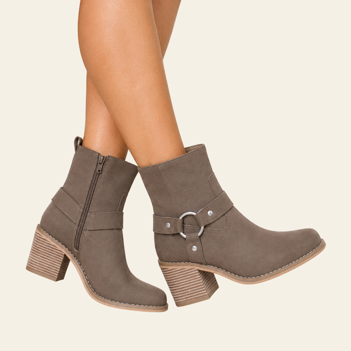 A pair of taupe-brown suede ankle boots with stacked block heels, side zippers, and decorative buckle straps, shown on a model's feet against a light cream background.