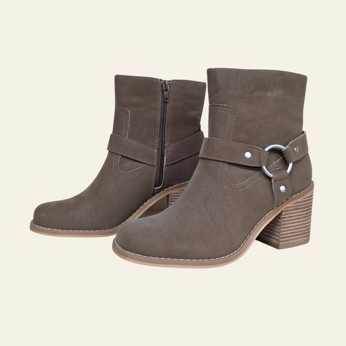 A pair of taupe-brown suede ankle boots with side zippers, decorative straps, and stacked block heels, shown against a solid cream background.