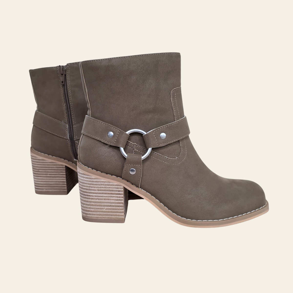 A pair of taupe-brown suede ankle boots with a block heel, side zipper, and a decorative strap with a silver-tone ring detail on the side, shown against a solid cream background.