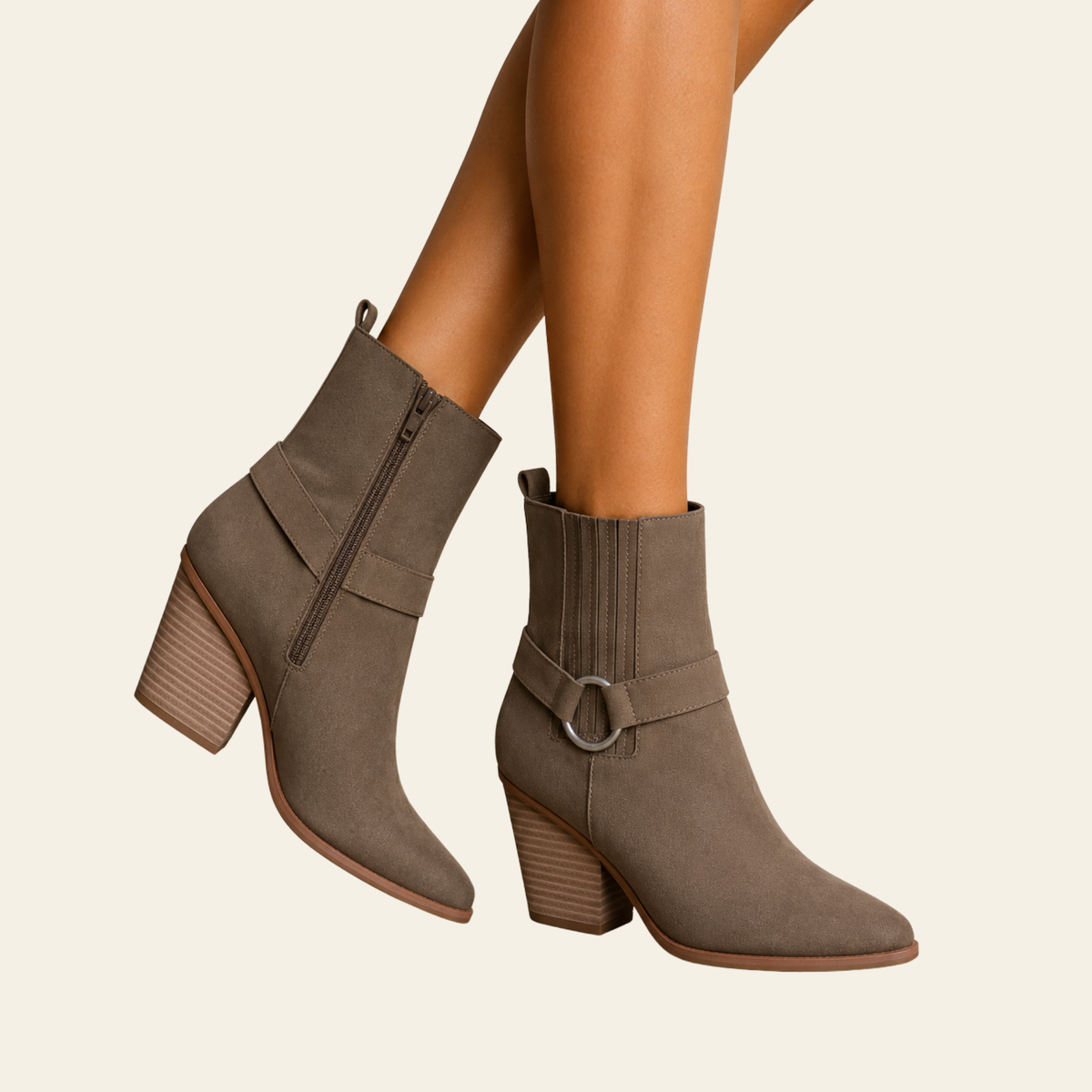 Taupe brown suede ankle boots with block heels, shown on a model's feet against a light cream background. One boot features a side zipper, and the other has a decorative strap with a silver-tone ring buckle.