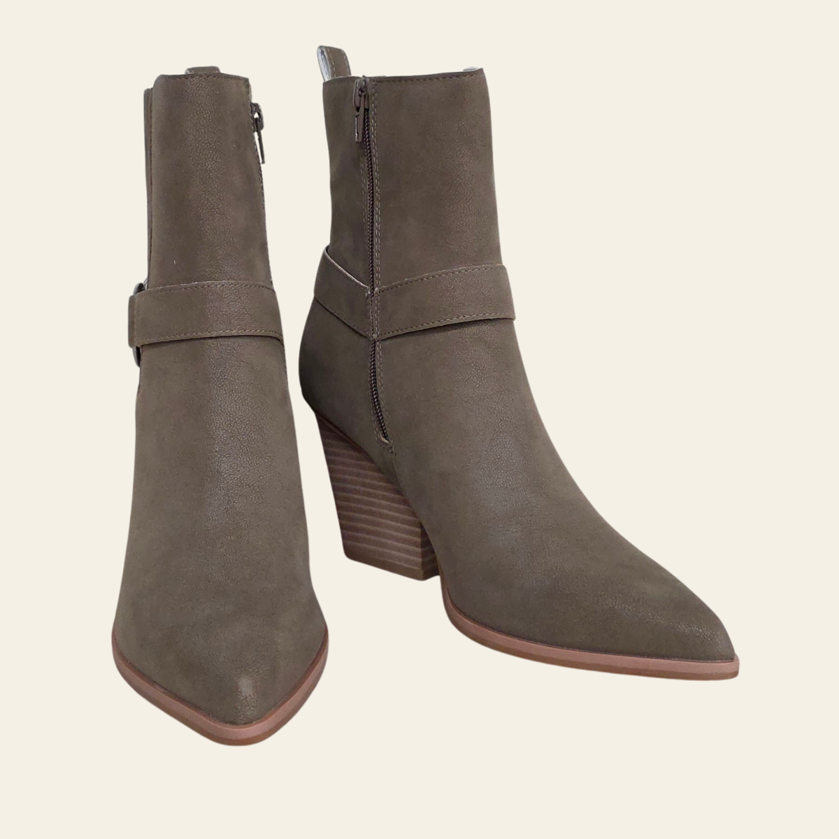A pair of taupe-brown suede ankle boots with pointed toes, low stacked block heels, and side buckle straps, shown against a solid cream background.