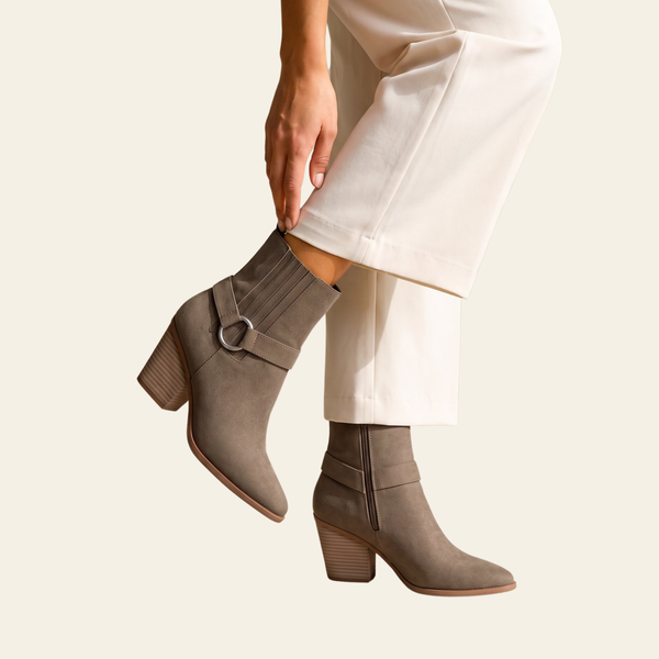 A person wearing taupe-brown ankle boots with a side buckle strap and block heel, paired with off-white cropped pants, against a solid light beige background.