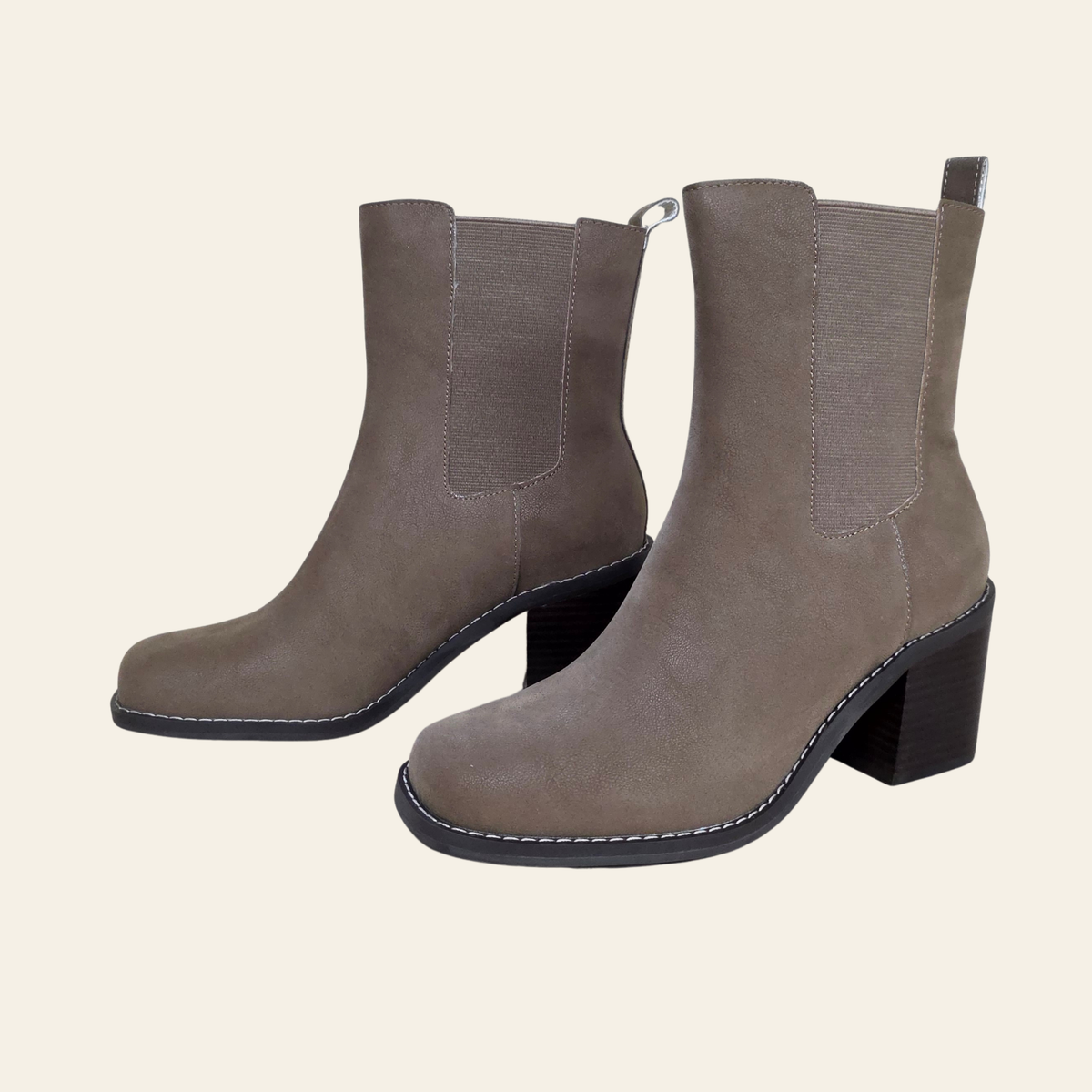 A pair of taupe-brown faux suede Chelsea-style ankle boots with black block heels and black stitching, shown against a solid cream background.