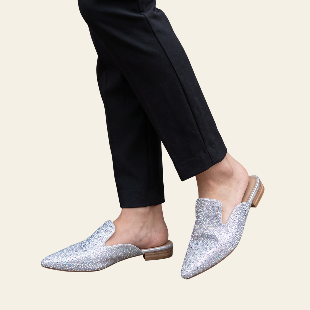 A person wearing pointed-toe slip-on mule flats covered in rhinestones paired with black pants, shown against a plain light beige background.