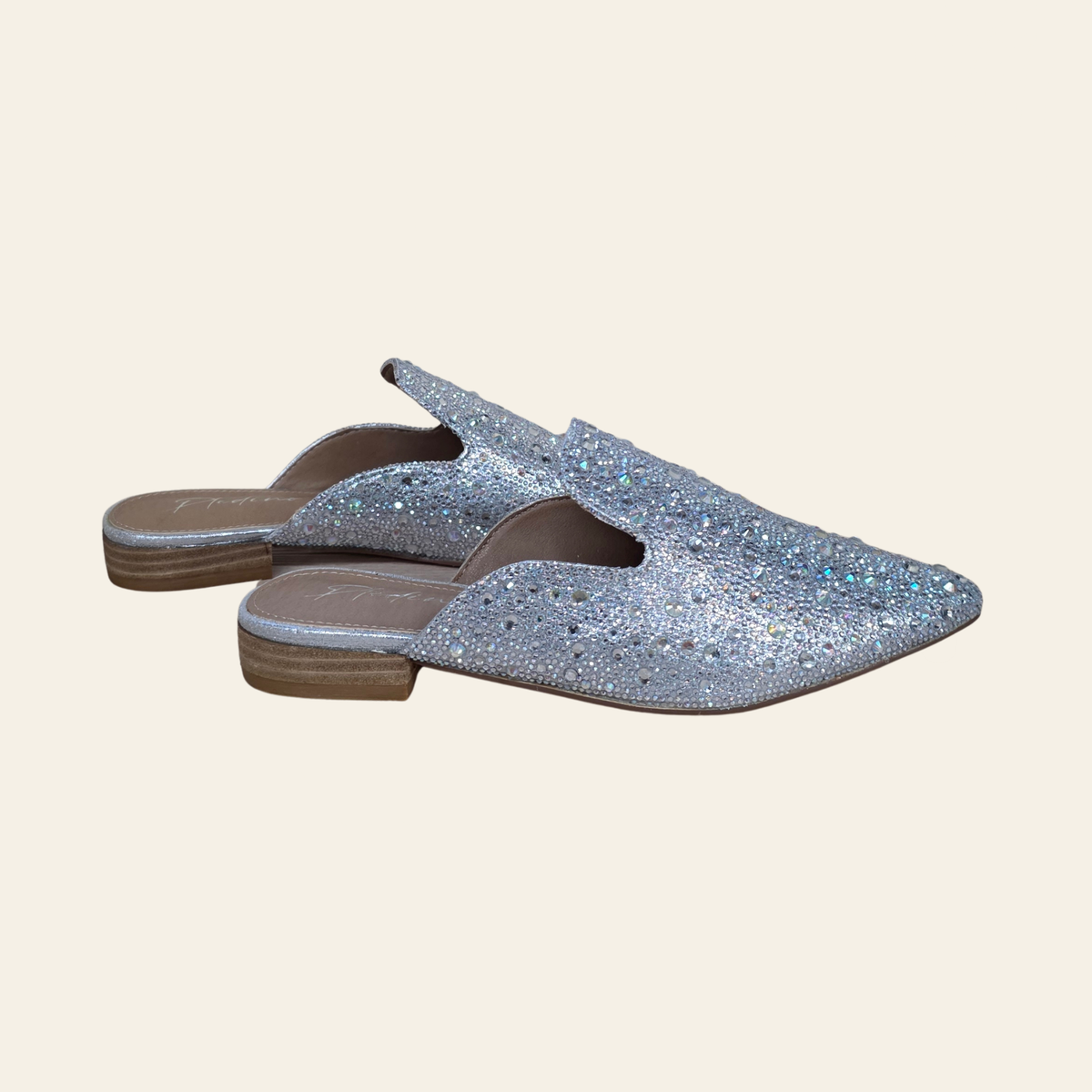 A pair of open-toe mule flats covered in rhintestones, featuring a low block heel and a brown sole, shown against a solid light beige background.
