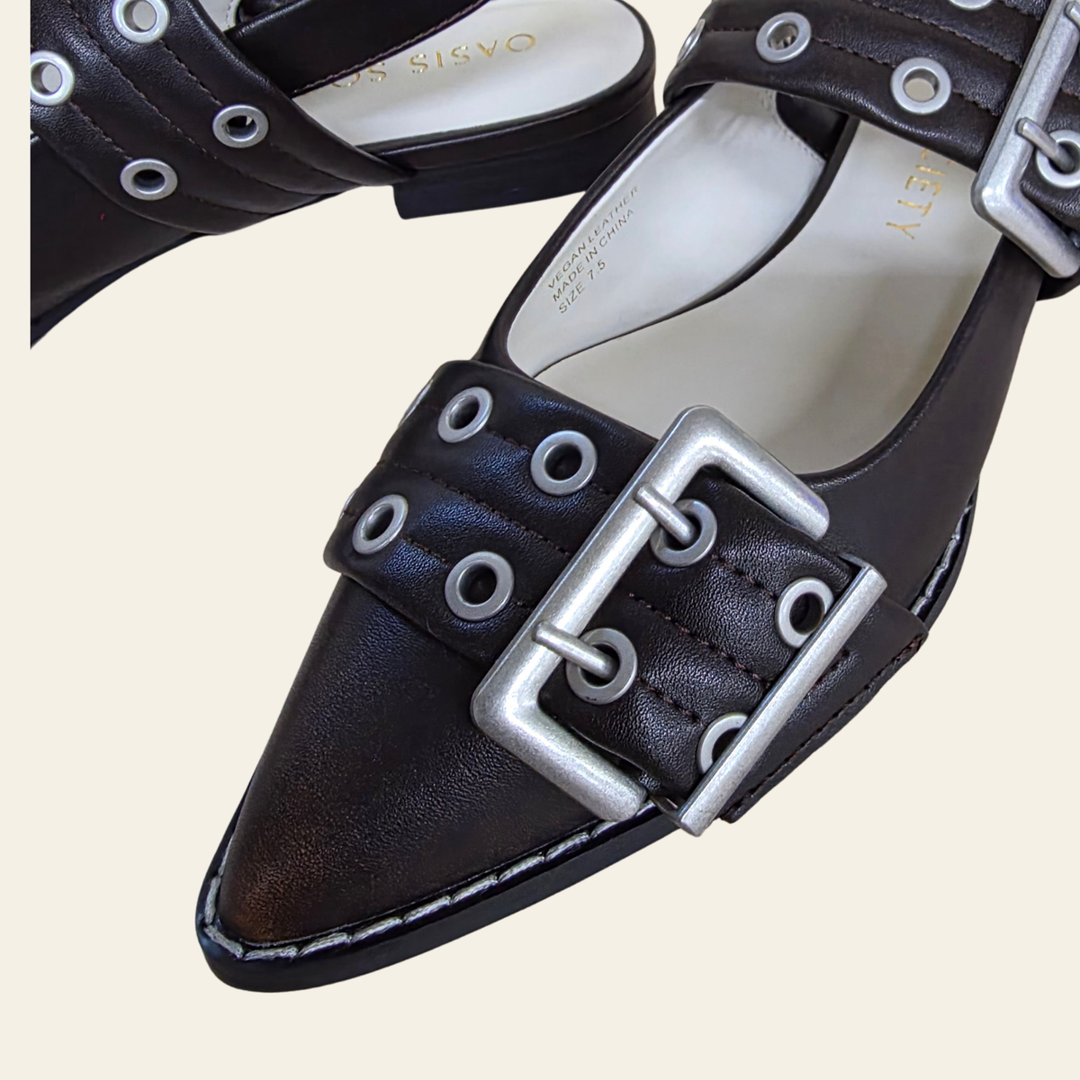 Brown leather pointed-toe flats with silver-tone metal grommets and large buckle straps, shown on a light beige background. The insole displays the brand name 'OASIS WEAR'.