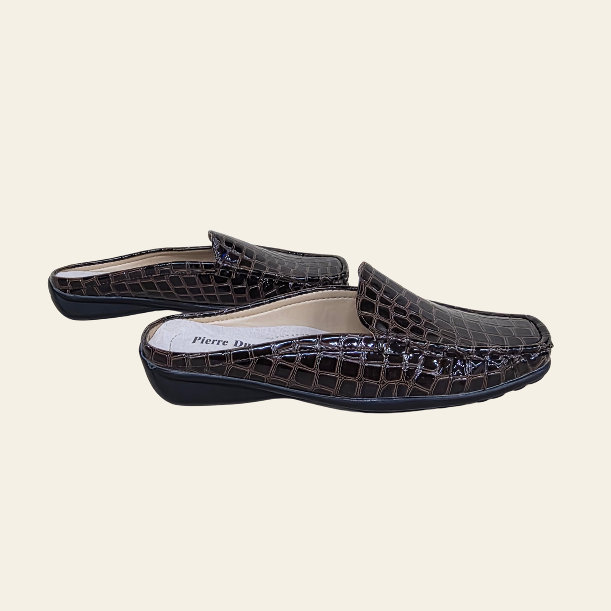 A pair of dark brown crocodile-embossed slip-on mule shoes with black soles, displayed against a solid light beige background. The brand name 'Pierre Dumas' is visible on the insole.