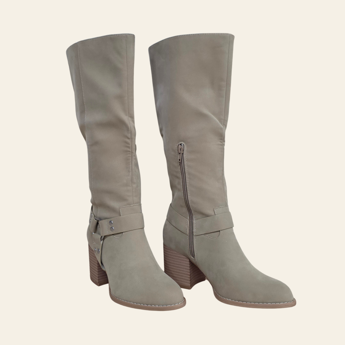 A pair of knee-high taupe suede boots with a side zipper, a decorative buckle strap at the ankle, and a stacked block heel, shown against a solid cream background.