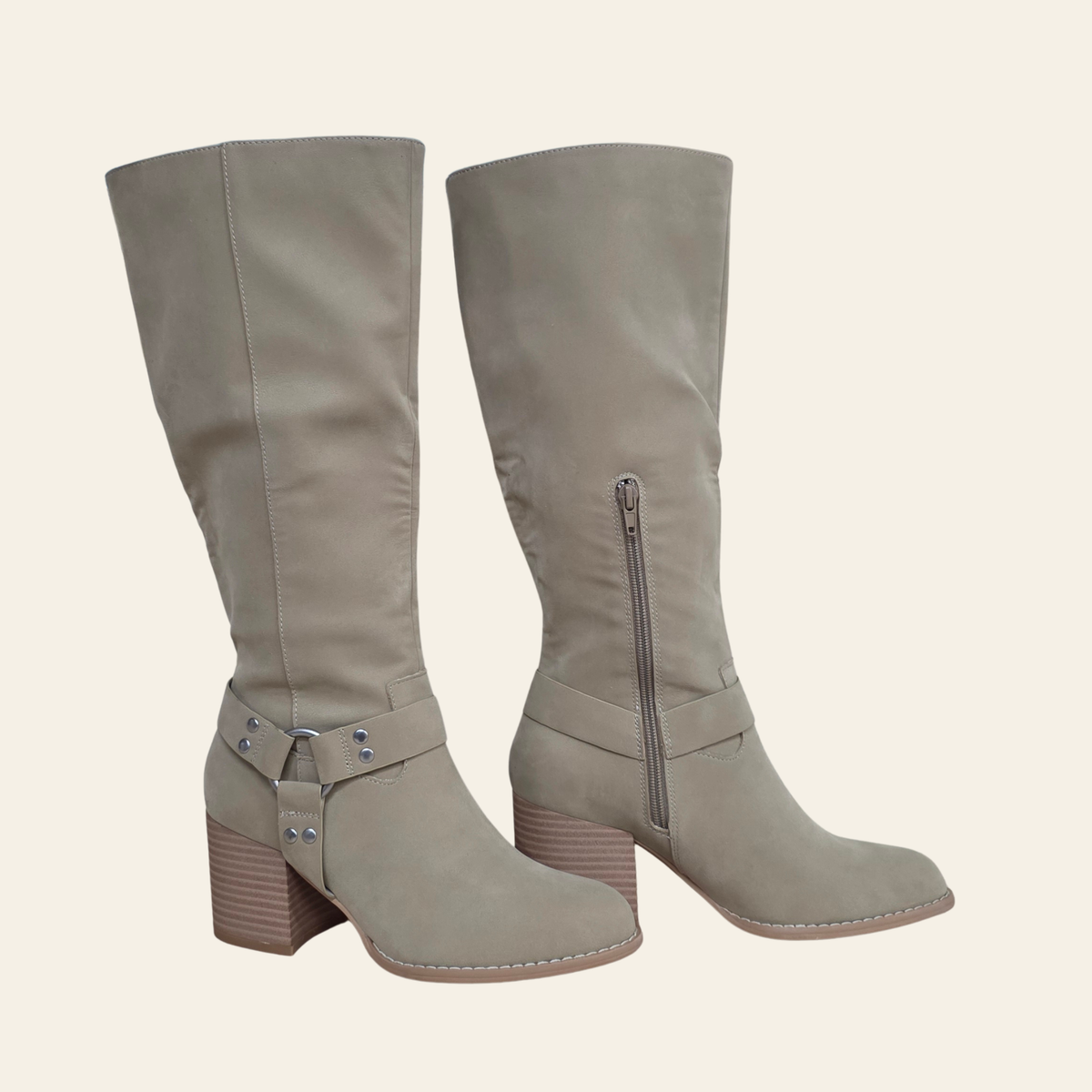A pair of knee-high taupe faux suede boots with block heels, side zippers, and buckle strap details on the lower shaft, shown against a solid cream background.