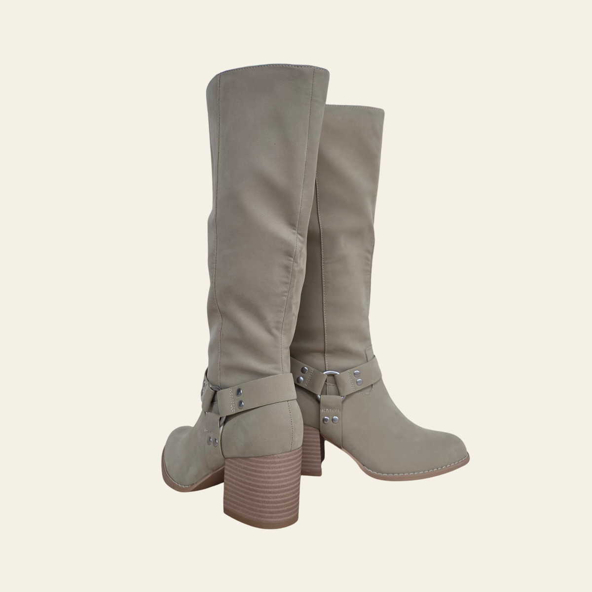 A pair of knee-high taupe suede boots with buckle straps and stacked block heels, shown from a three-quarters angle against a solid cream background.