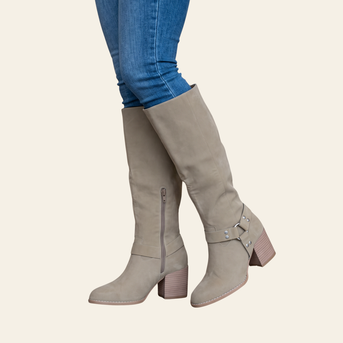 Taupe knee-high boots with harness straps, silver stud accents, partial side zippers and stacked block heels, styled with blue jeans.