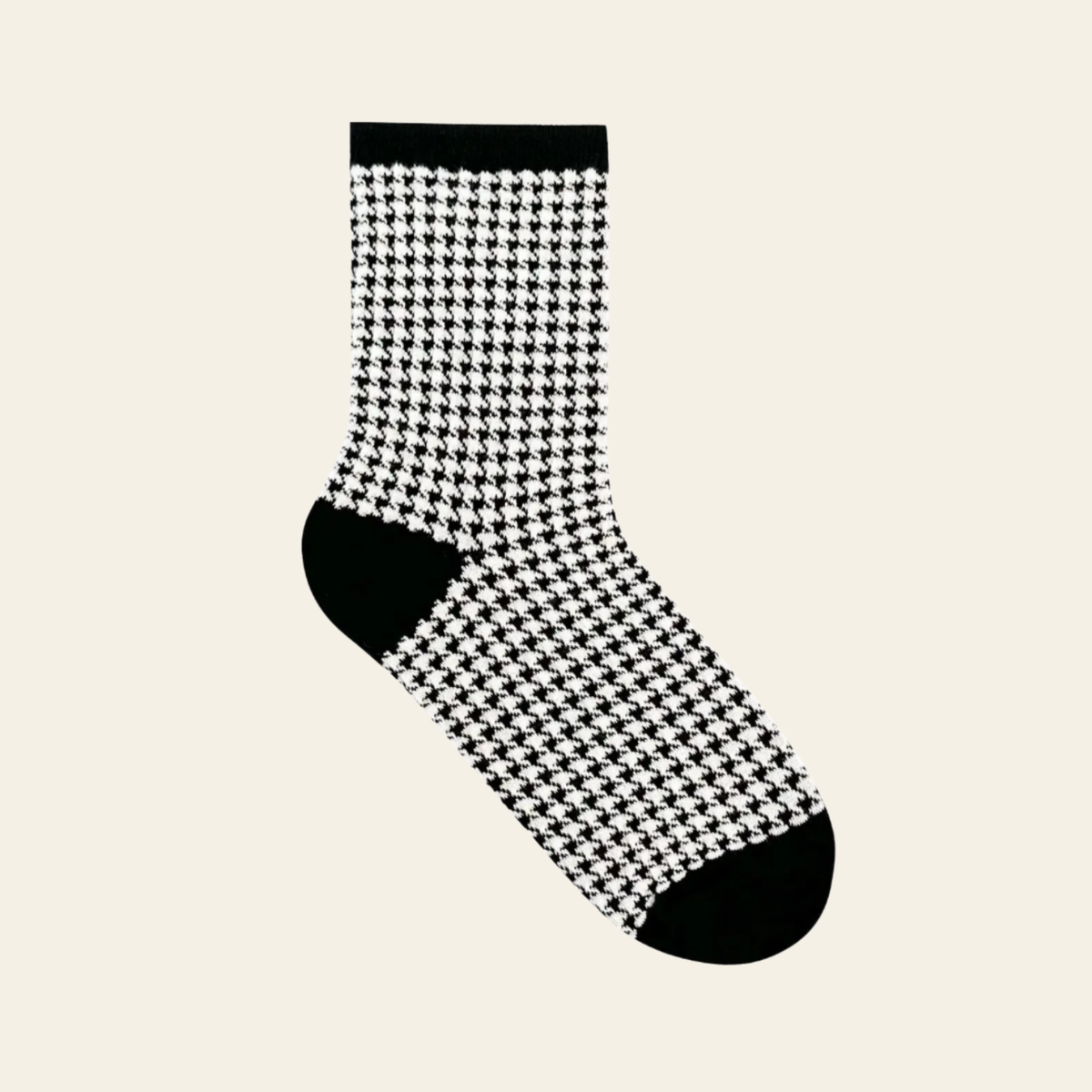 A single crew sock with a black and white houndstooth pattern, featuring solid black heels, toes, and a ribbed cuff, shown against a solid light beige background.