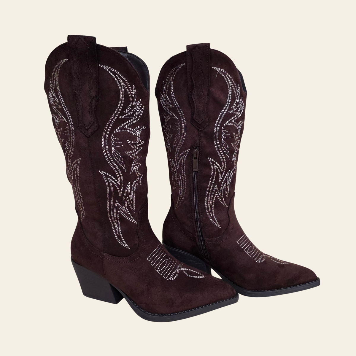 A pair of dark brown suede cowboy boots with white western embroidery and rhinestone detailing, shown against a solid light beige background.