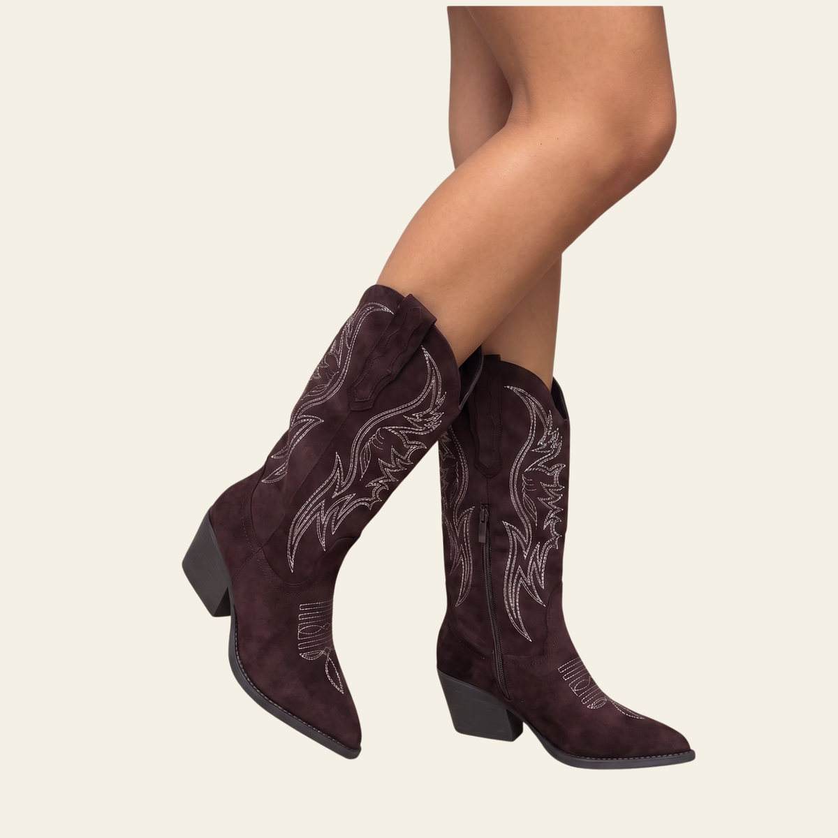 A pair of dark brown suede cowboy boots with white embroidered western patterns, shown on a model's legs against a light beige background.