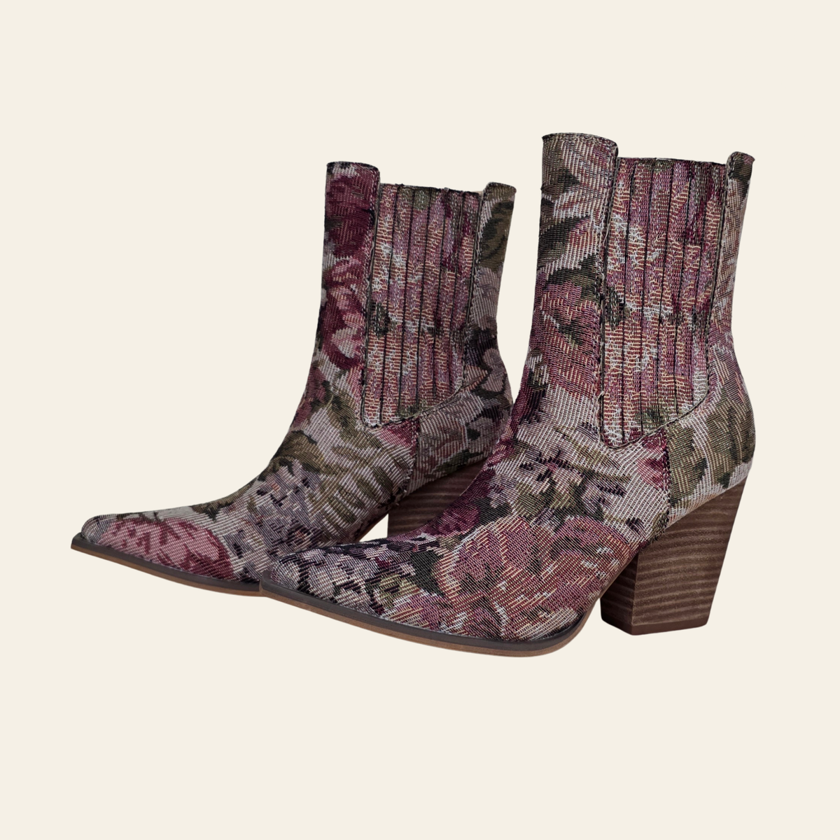 A pair of pointed-toe ankle boots with a woven floral tapestry pattern in dusty pink, mauve, and olive green, featuring a ribbed elastic side gore and a brown block heel, set against a solid cream background.