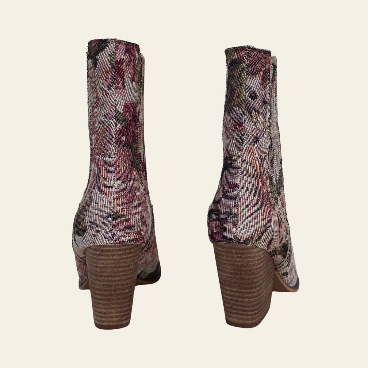 A pair of women's ankle boots shown from the back, featuring a textured floral tapestry pattern in muted pink, purple, and green tones, paired with chunky brown stacked block heels, set against a solid light beige background.