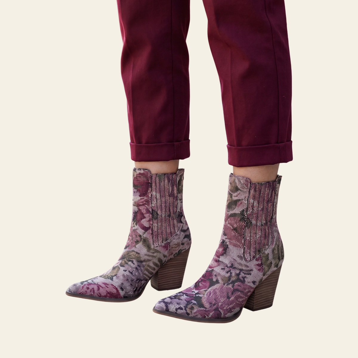 A pair of pointed-toe ankle boots with a pink and purple floral pattern, worn with dark burgundy pants against a light cream background.