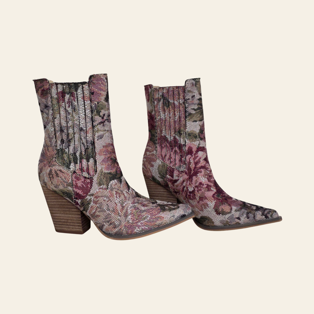 A pair of pointed-toe ankle boots with a textured floral tapestry pattern in muted pink, burgundy, and olive green, featuring a stacked wood-look block heel, set against a solid cream background.