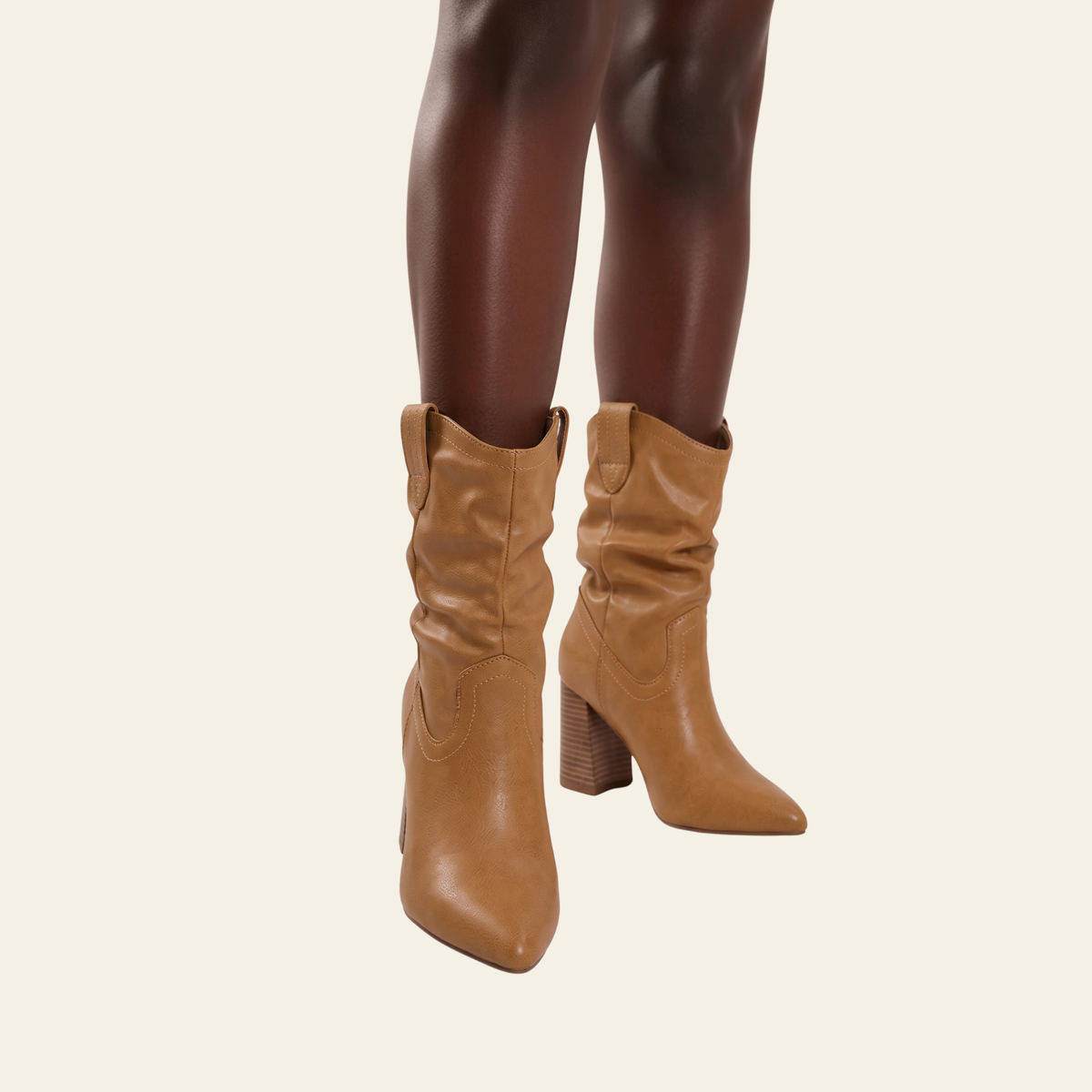 Tan faux leather slouchy ankle boots with a block heel and pointed toe, shown on a model's legs against a light cream background.