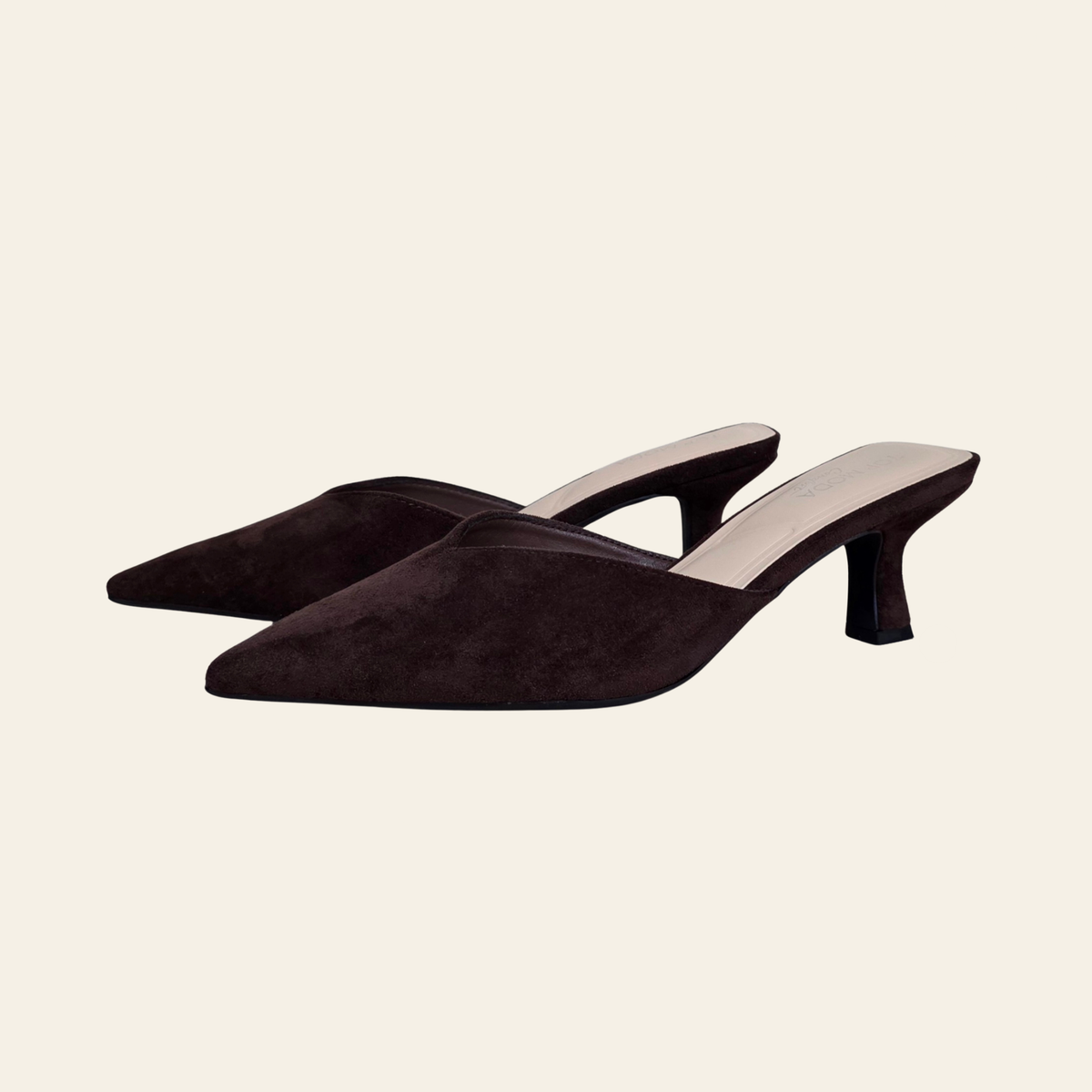 A pair of dark brown suede mule heels with pointed toes and low block heels, displayed on a light cream background.