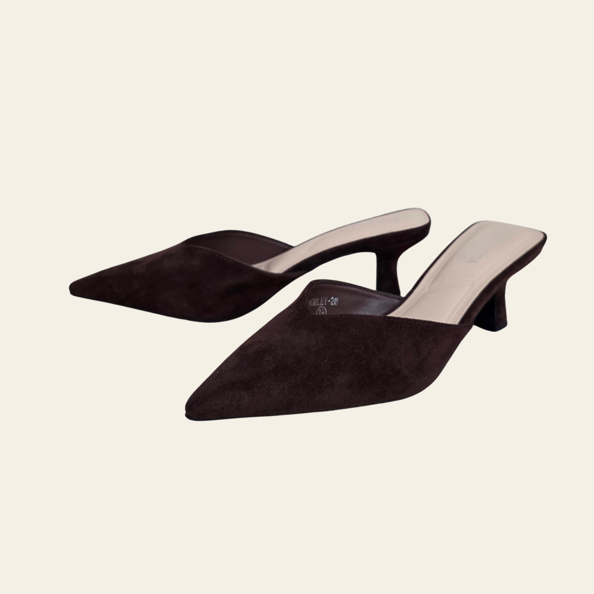 A pair of dark brown suede pointed-toe mule heels with a low block heel, displayed on a solid light beige background.
