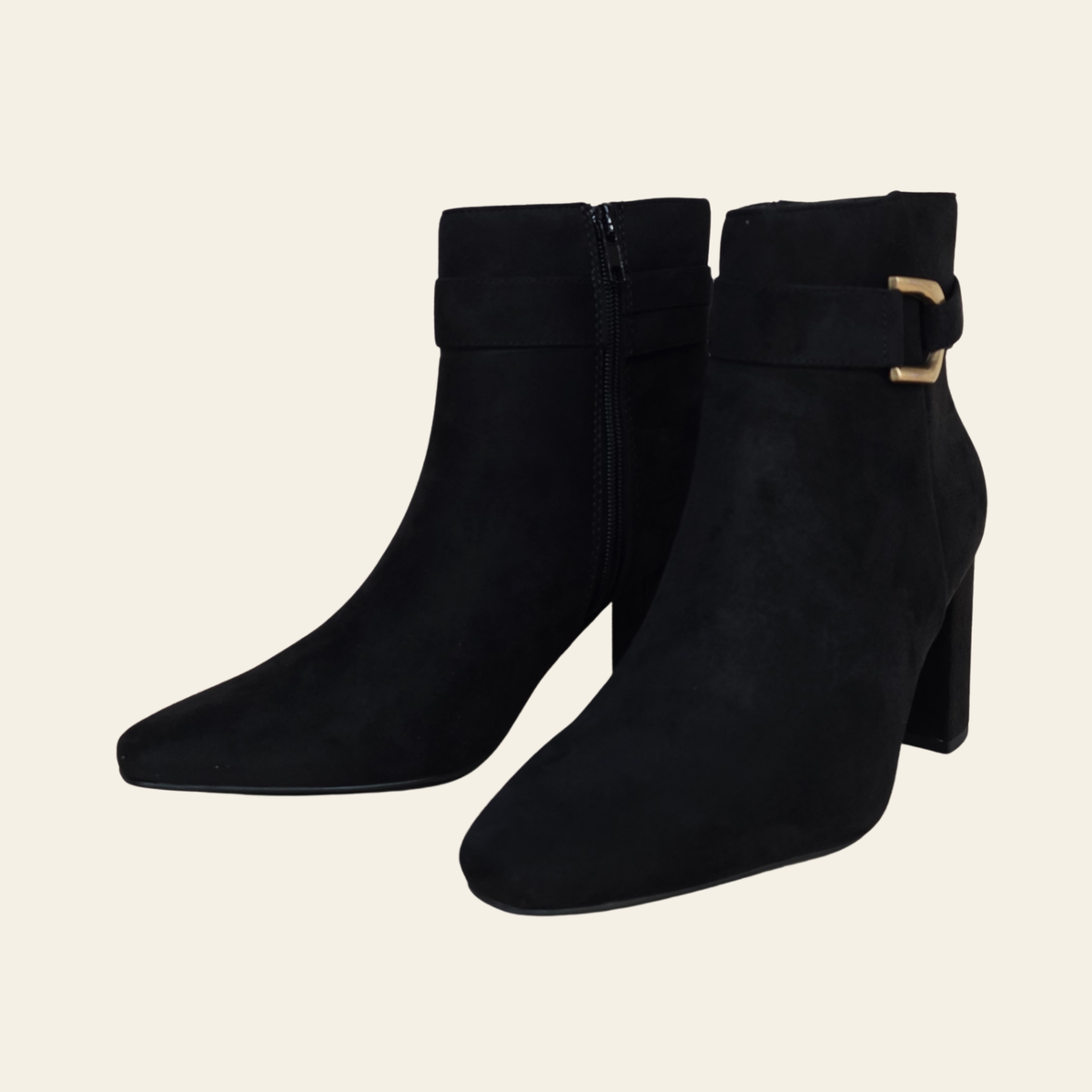 A pair of black faux suede ankle boots with block heels, side zippers, and decorative buckle straps on the outer side, shown against a solid light beige background.
