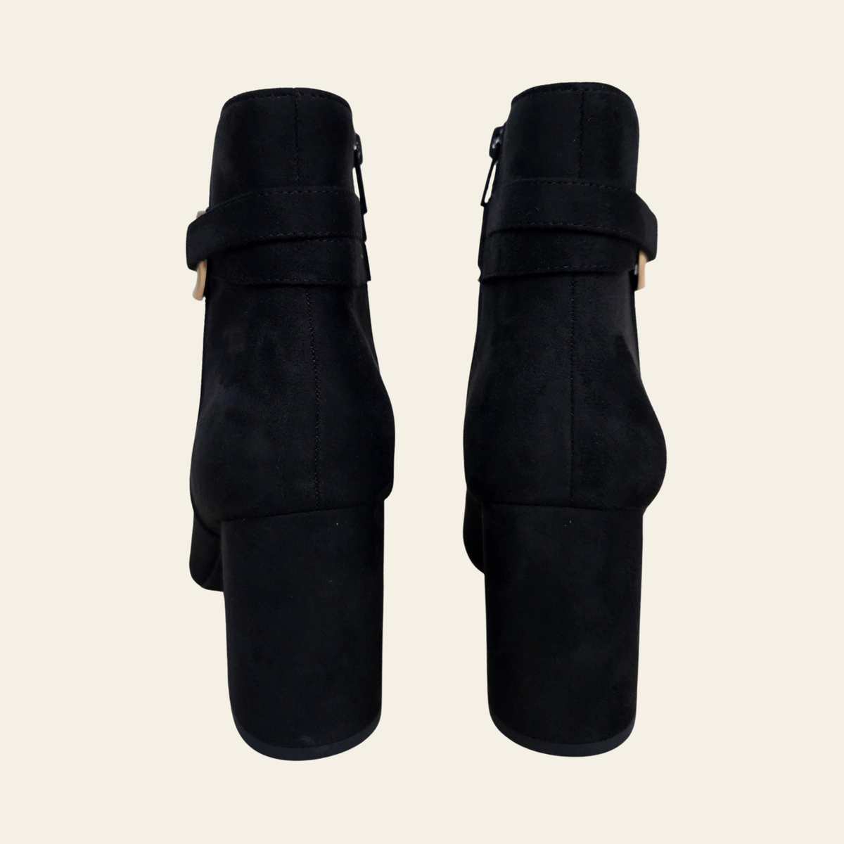 A pair of black faux suede ankle boots with block heels and a wide strap detail with a small gold-tone buckle, shown from the back on a light beige background.
