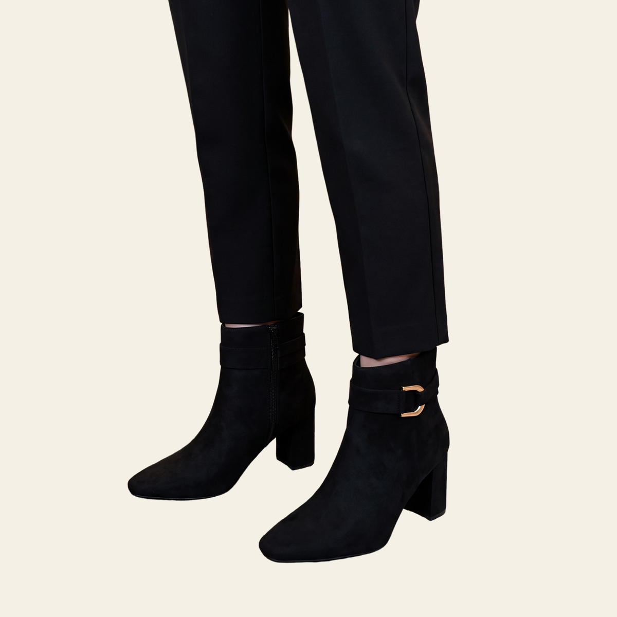 Black ankle boots with block heels and a gold buckle detail, shown on a model wearing black trousers against a light beige background.