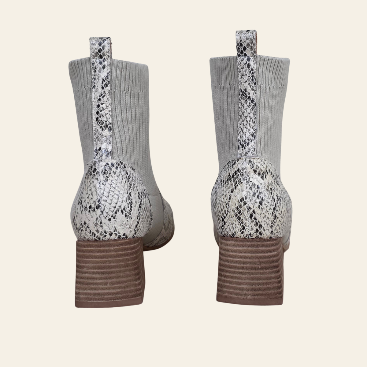 A pair of women's ankle boots shown from the back, featuring gray ribbed knit socks, a white and black snakeskin pattern on the heel and pull tabs, and brown stacked block heels, set against a solid cream background.