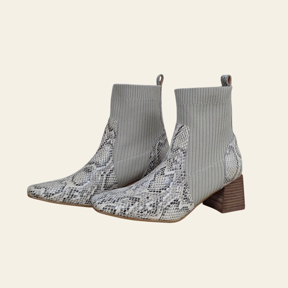 A pair of pointed-toe ankle boots featuring a gray and black snakeskin pattern on the toe and heel, with ribbed gray knit uppers and brown block heels, set against a solid cream background.