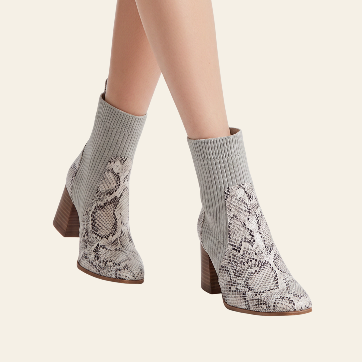 A pair of women's ankle boots with a gray ribbed knit sock-like upper and a beige and gray snakeskin pattern on the lower portion, shown on a model against a light cream background.