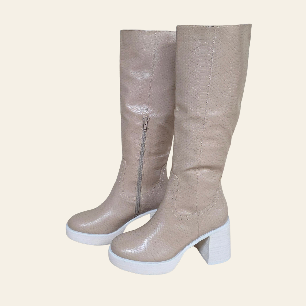 A pair of knee-high boots in a light beige faux-leather material with a subtle textured pattern, featuring a white platform sole and a chunky white block heel. One boot is shown from the side, revealing a silver zipper, while the other is shown from a three-quarter angle.