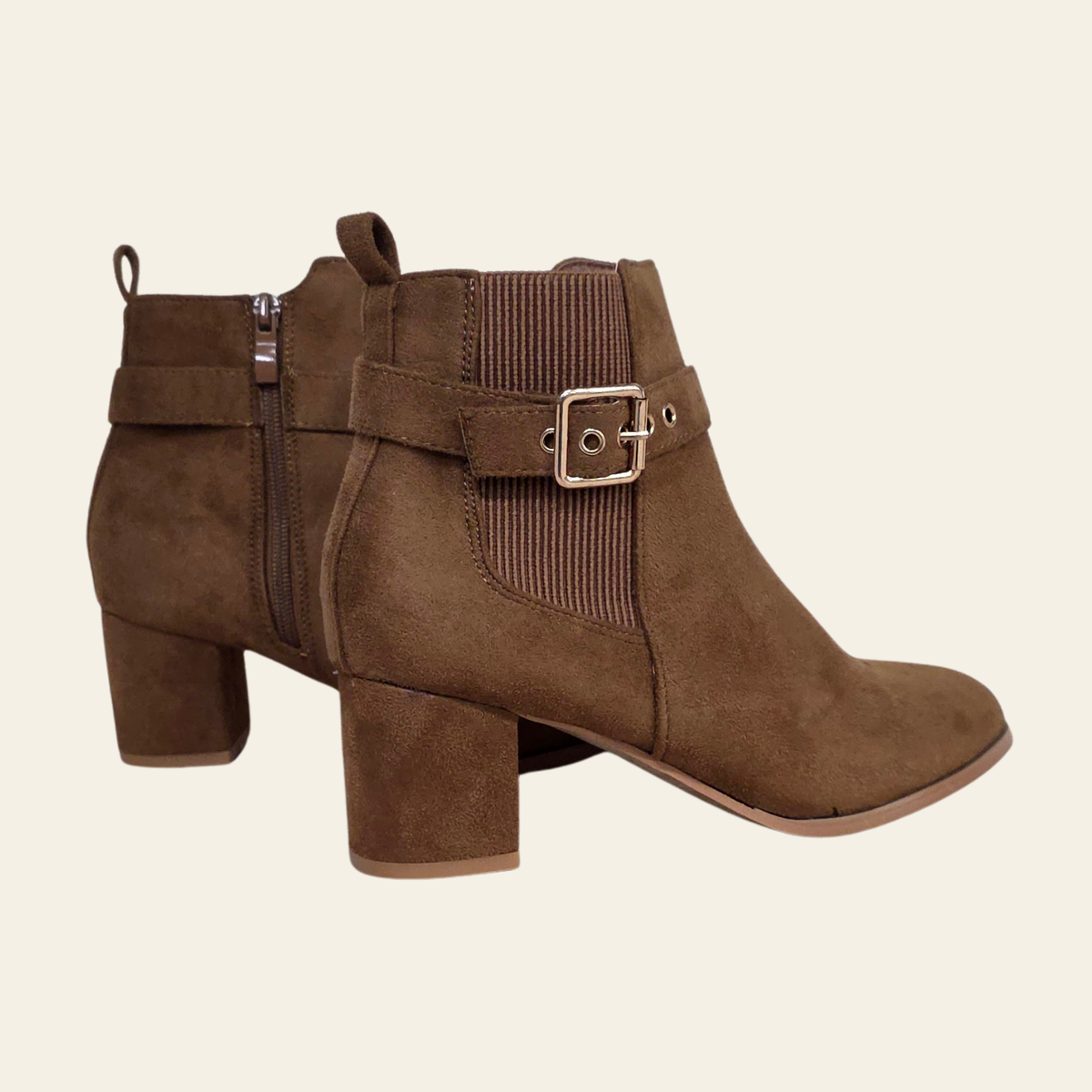 A pair of women's brown suede ankle boots with a block heel, a side zipper, and a decorative elastic side strap with a metal buckle, shown against a solid light beige background.