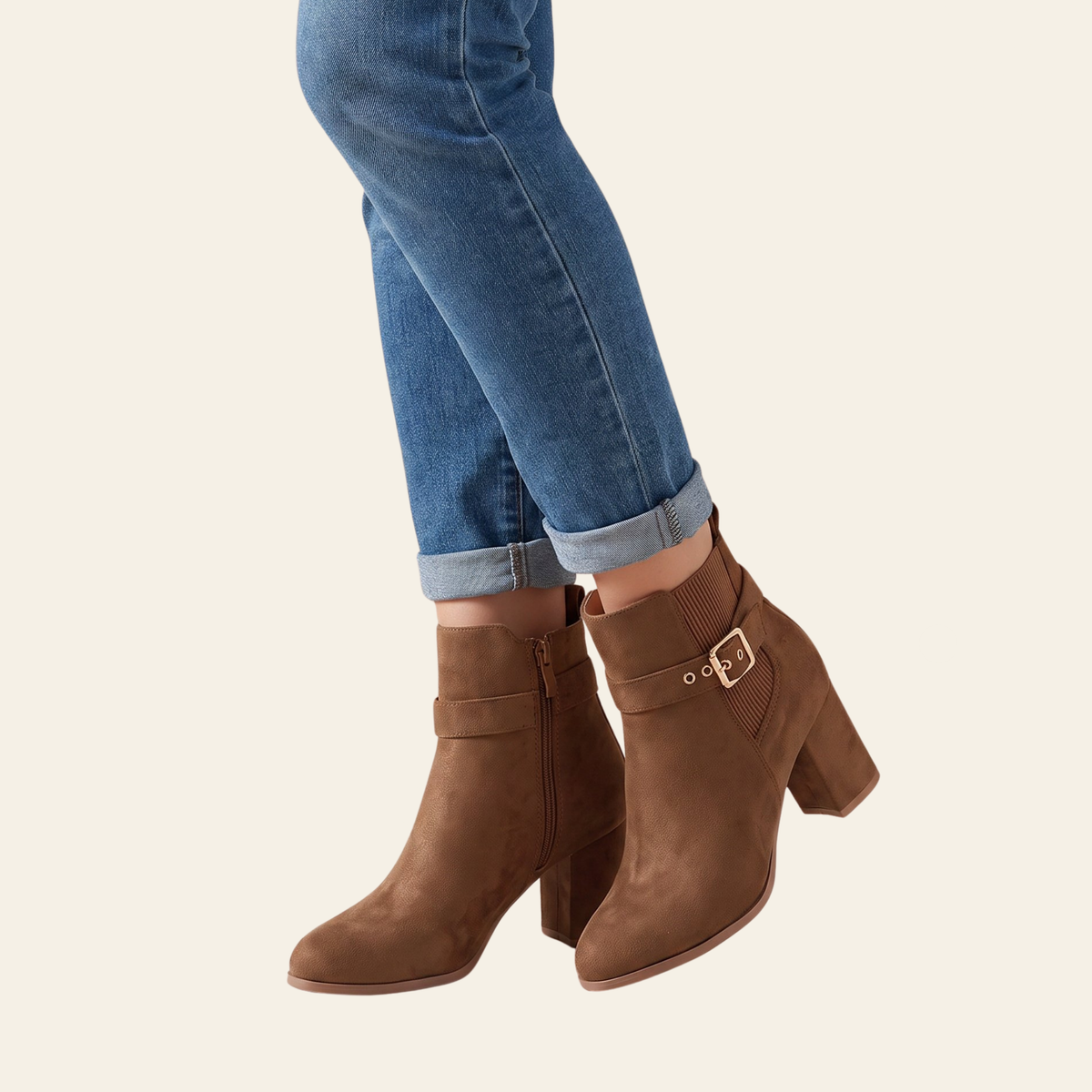 A pair of women's brown suede ankle boots with block heels and side buckle details, shown on a model wearing blue jeans against a light cream background.