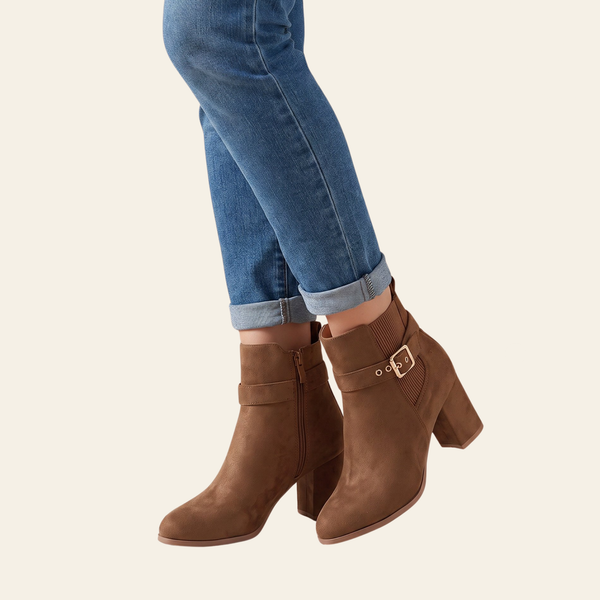 A pair of women's brown suede ankle boots with block heels and side buckle details, shown on a model wearing blue jeans against a light cream background.