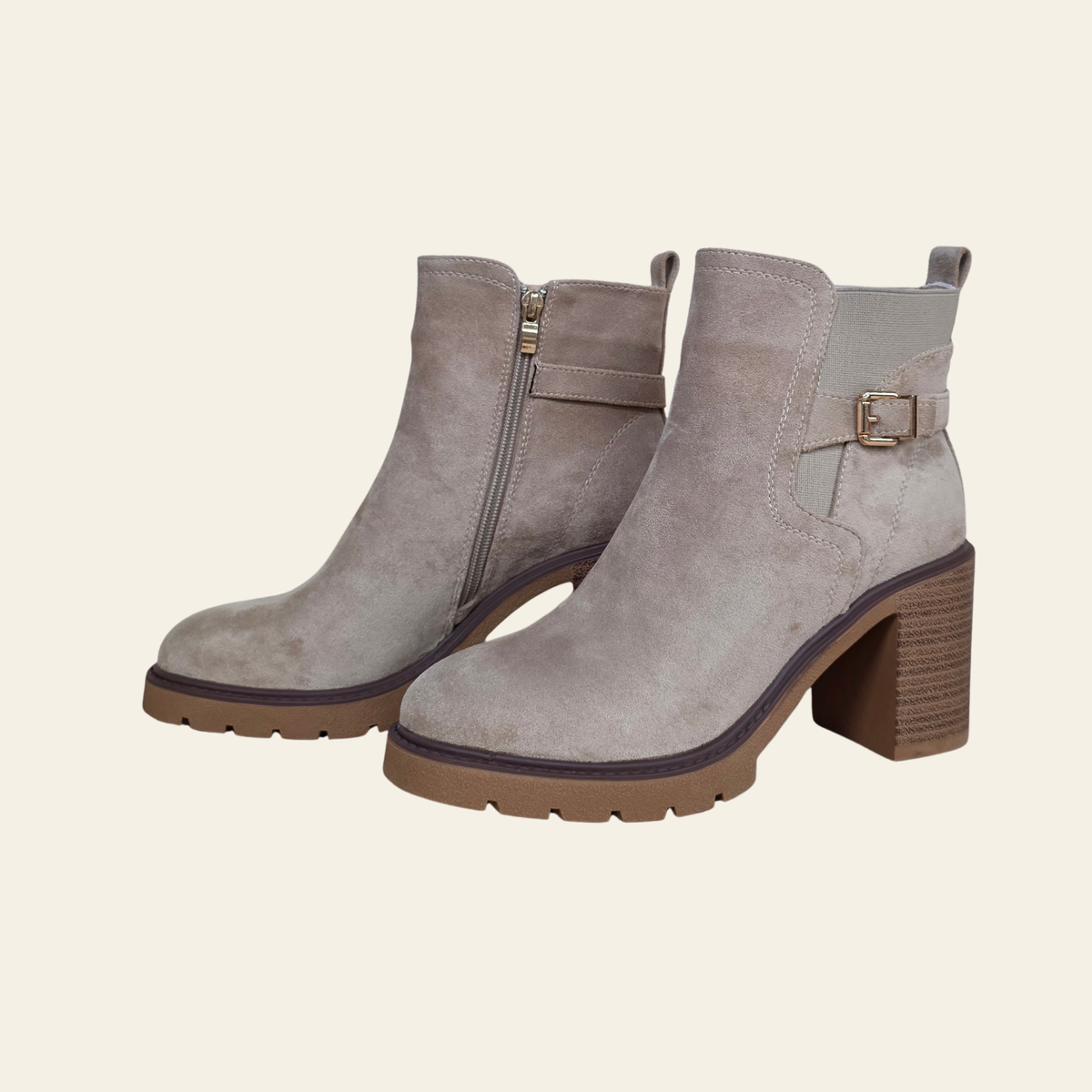 A pair of taupe suede ankle boots with block heels, featuring a side zipper on one boot and a decorative buckle strap on the other, set against a solid cream background.