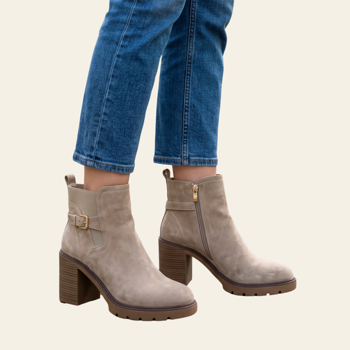 Taupe suede ankle boots with a block heel, side buckle strap, and side zipper, shown on a model wearing blue jeans against a light beige background.