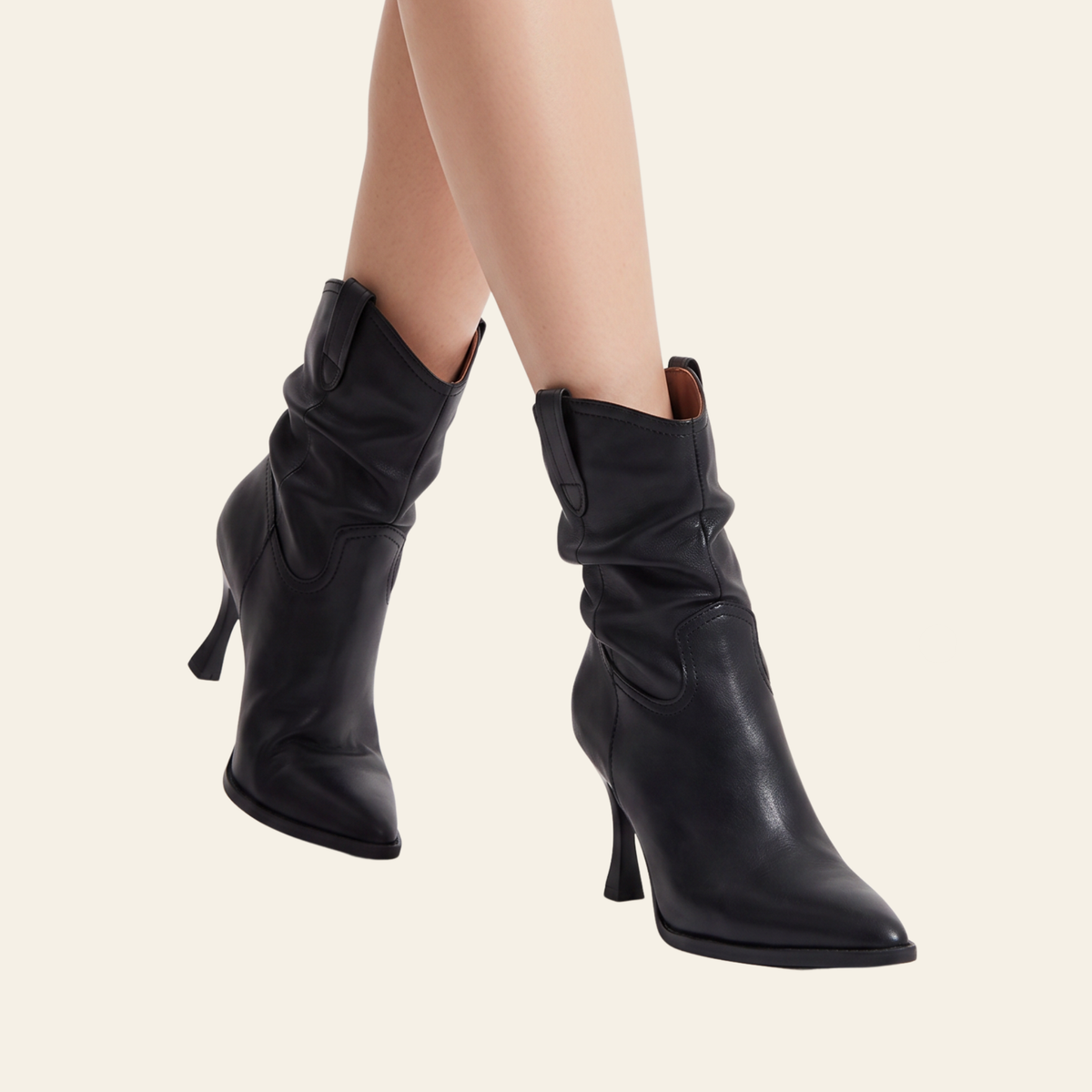 A pair of black leather slouchy ankle boots with pointed toes and mid-height stiletto heels, shown on a model's feet against a light cream background.