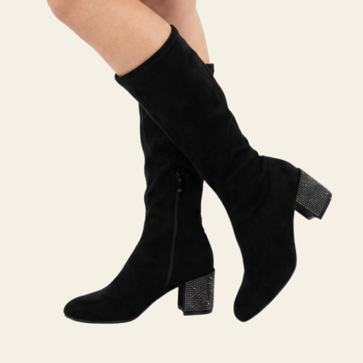 Black suede knee-high boots with block heels and rhinestone-embellished heels, shown on a model's legs against a light beige background.