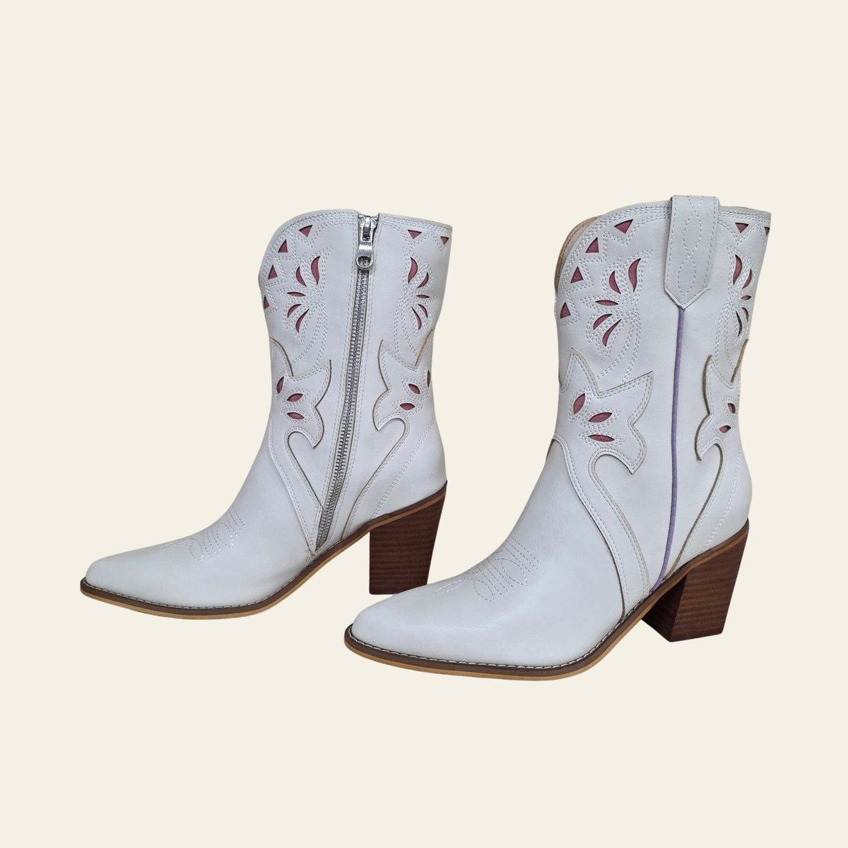 A pair of white leather cowboy boots with brown block heels, featuring decorative cutouts and western-style stitching on the shafts, shown against a solid light beige background.