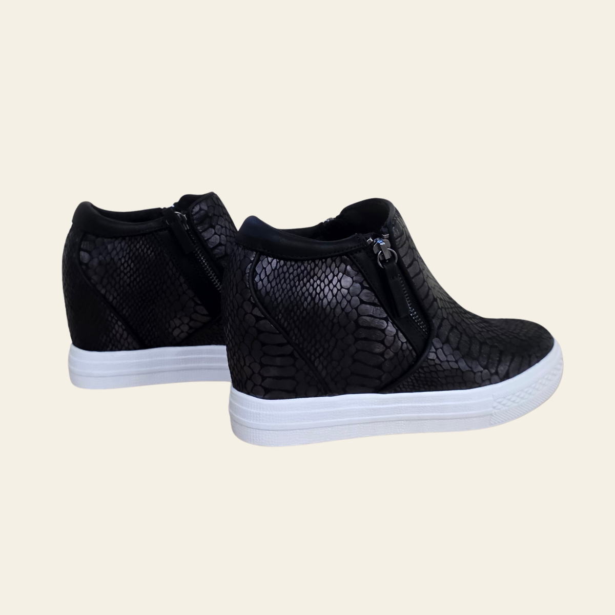 A pair of black wedge sneakers with a snakeskin texture, featuring side zippers and thick white platform soles, shown against a solid light beige background.