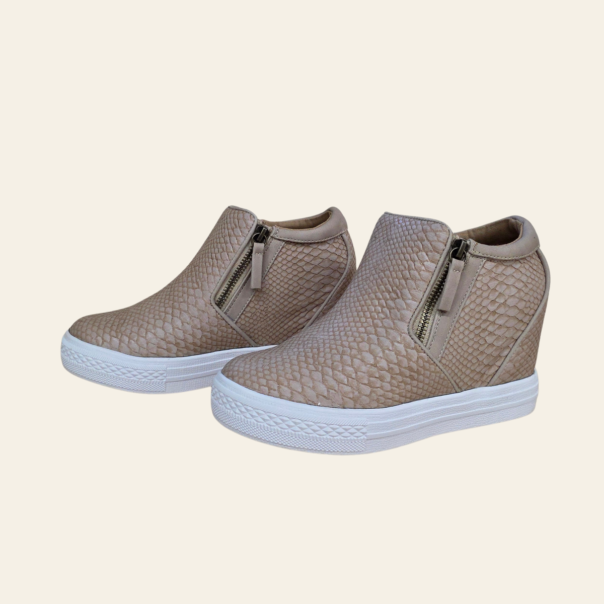 A pair of women's wedge sneakers in a textured snakeskin pattern, featuring a light blush, side zippers, and a white platform sole, set against a solid cream background.