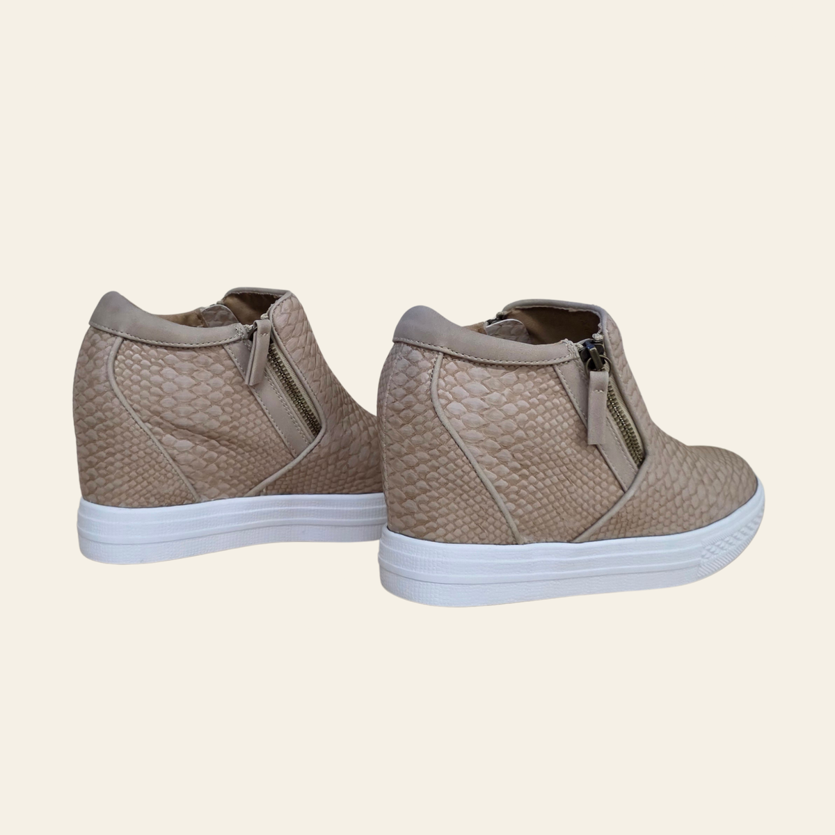 A pair of women's wedge sneakers in a textured blush faux-snakeskin pattern, featuring side zippers and thick white platform soles, shown against a solid cream background.