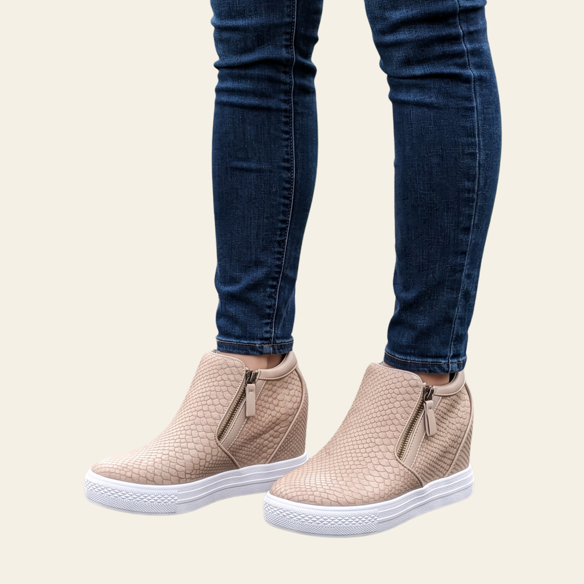 A person wearing blush textured ankle slip-on sneakers with white soles and dark blue skinny jeans, shown from the knees down against a light cream background.