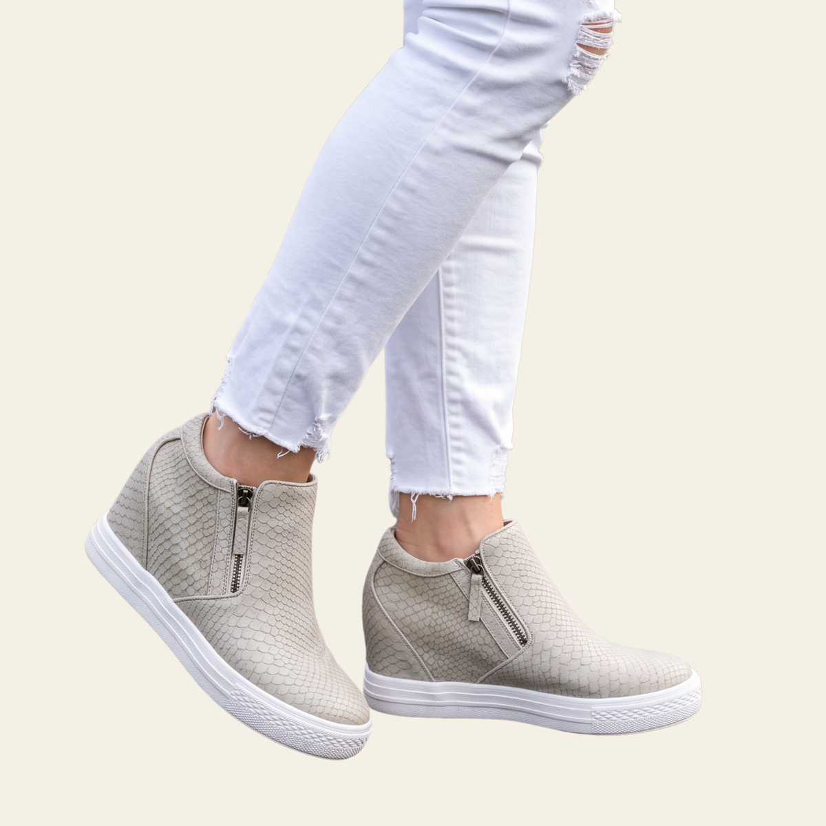A pair of women's cream colored, textured slip-on sneakers with side zippers, shown on a model wearing white ripped jeans against a light beige background.