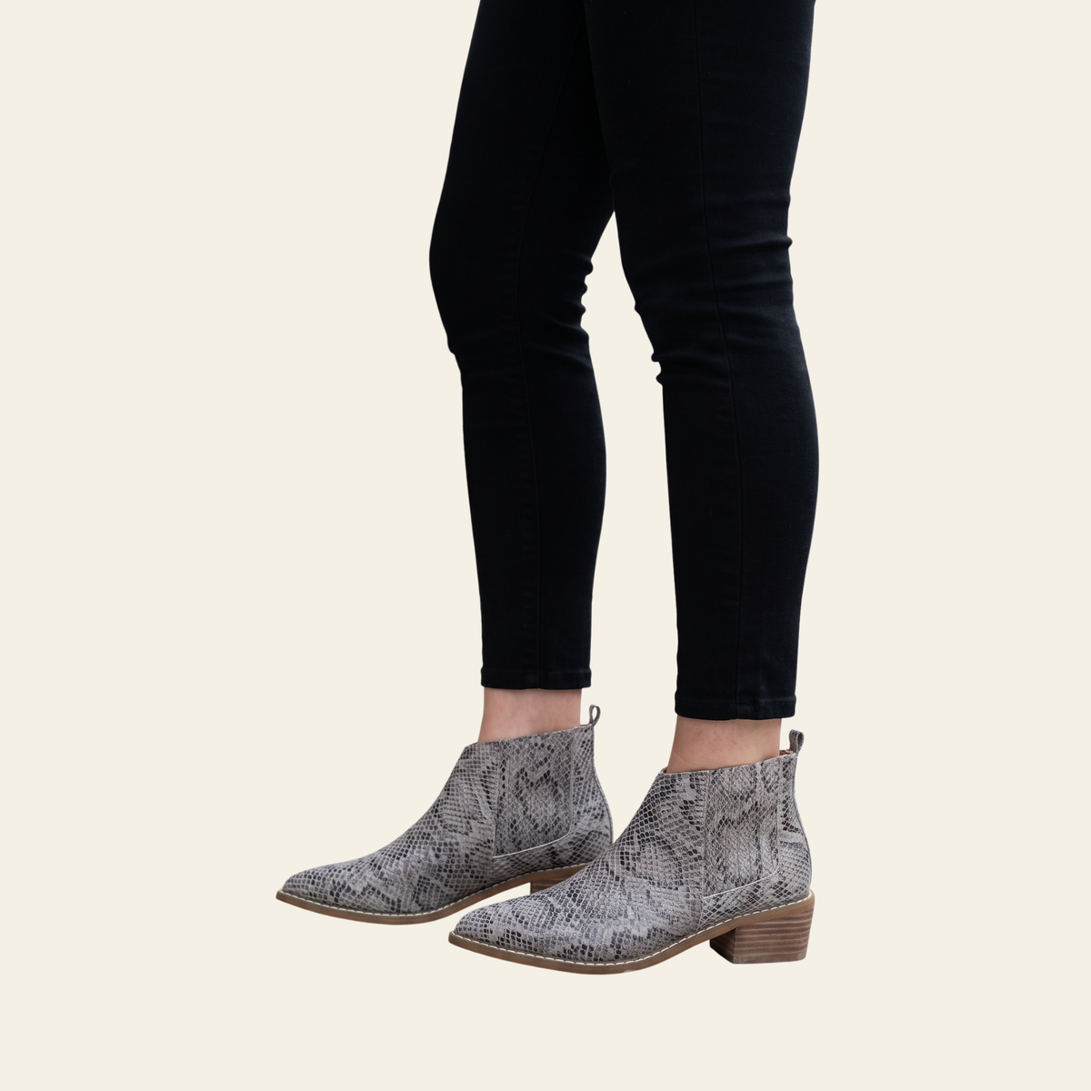 Gray and black snakeskin-patterned ankle boots with low block heels, shown on a model wearing black skinny jeans against a light cream background.